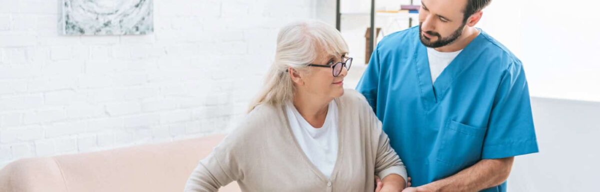 A healthcare worker in blue scrubs supports an elderly woman with white hair and glasses as she uses a cane, highlighting compassion beyond traditional views of masculinity and gender identity in caregiving roles.