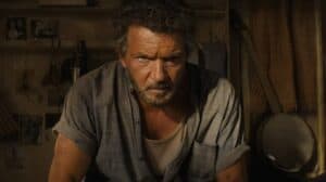 A man with graying hair and a serious expression leans forward in a dimly lit room, the banjo and photos on the wall behind him evoking stories of spiritual growth and reflection, much like the Parable of the Soil—a reminder of cultivating a life that is capable of producing fruit.