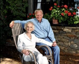 An elderly couple enjoys each other's company outdoors, holding hands as the woman sits in a chair and the man on the armrest. With a stone wall and red flowers behind them, they share quiet blessings within their community.