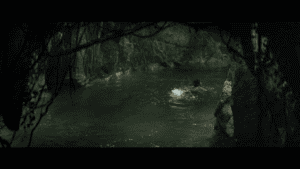 A person swims in a small, rocky, forested pool surrounded by overhanging roots and foliage, embodying the parable of the soil—a quiet reflection on spiritual growth amid nature's complexity.
