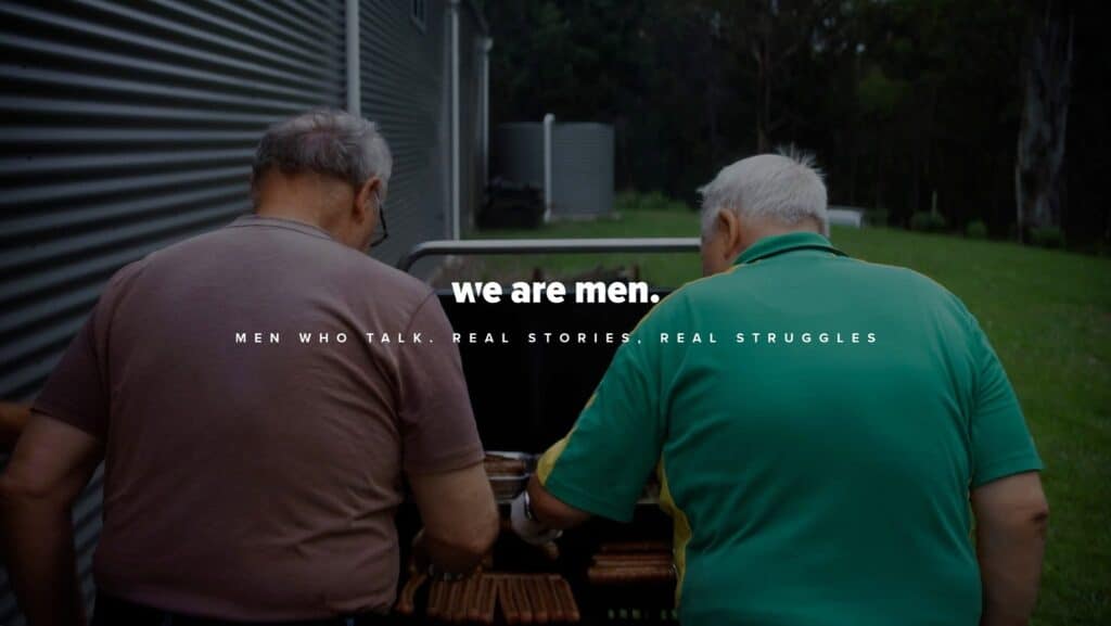 Two older men stand side by side at a barbecue grill outdoors, sharing their stories. Text overlay reads: "we are men. Men who talk. Real stories. Real struggles." Their candid conversation highlights the impact of their projects and media appearances, inviting others to join in honest dialogue.