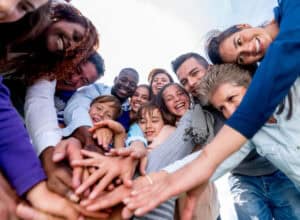 A diverse community of people standing in a circle outdoors, smiling and stacking their hands together in the center, enjoying the moment.