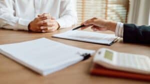 Two people sit at a desk with documents, a clipboard, and a calculator; one person gestures with a pen toward the paperwork as they discuss community blessings.