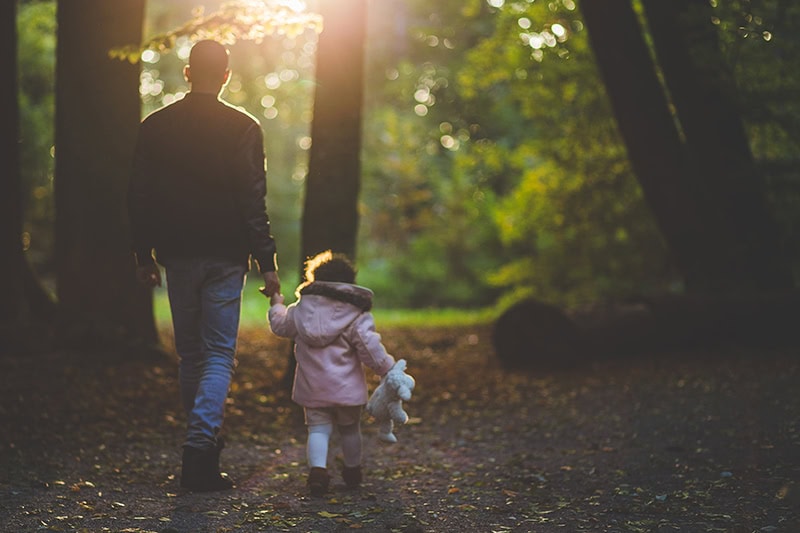 An adult and a child holding hands walk through a sunlit forest path, embodying the warmth and hope of Christian living. The child, wearing a pink coat and holding a stuffed animal, reflects the simple joys found in following Jesus’ teachings.