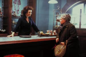 A woman stands behind a counter speaking to an older woman holding a round woven bag inside a shop with cakes displayed on a shelf, evoking the spirit of Feeding The 5000 as they share a moment of connection over fresh treats.