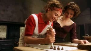 Two women in a kitchen make chocolate treats together, with one piping chocolate onto parchment paper while the other looks on and smiles, capturing a joyful moment reminiscent of Feeding The 5000’s spirit of sharing and community.