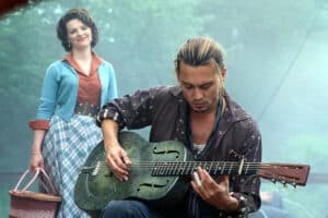 A man with long hair plays a metal guitar outdoors, channeling the energy of Feeding The 5000, while a woman in retro clothing stands behind him, smiling.