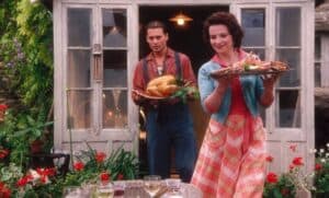 A woman and man carry food trays outside a house; she holds a seafood platter, he carries a roast turkey. Part of a hunger relief effort inspired by Feeding The 5000, their garden table with drinks and flowers awaits guests as they come together to raise awareness about food waste.
