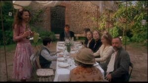Nine people sit around a table set for a meal in a garden under a canopy during a relaxed community event. One woman stands pouring a drink, as laughter and conversation fill the air. The scene has an informal, welcoming atmosphere reminiscent of Feeding The 5000, celebrating the joy of coming together over food while raising awareness about food waste.