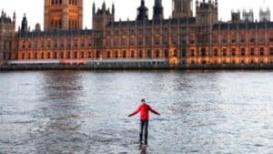 A person in a red jacket appears to walk on water on the surface of the River Thames, with the majestic Houses of Parliament standing in the background.
