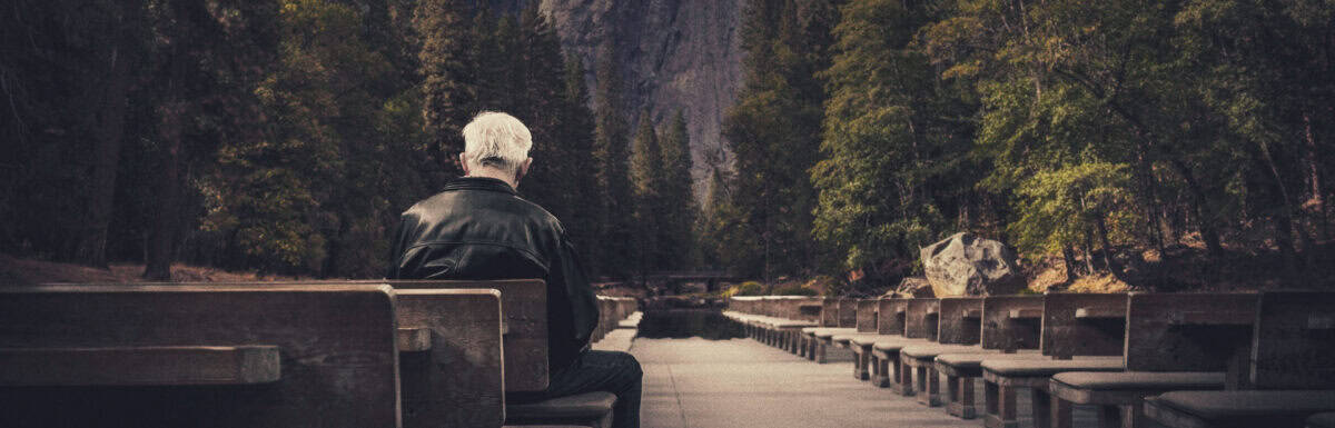 An older man with gray hair sits alone on a wooden bench outdoors, facing away, surrounded by empty benches and tall trees with mountains in the background, inviting quiet reflection on the motives behind his solitude and the psychology of loneliness.