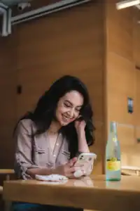 A woman sits at a table, smiling while looking at her phone. A bottle of sparkling water and a Walk On Water face mask are on the table in front of her.