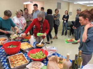 A group of people stand around a table covered with a blue floral cloth, serving themselves food from various dishes at a casual indoor gathering reminiscent of Feeding The 5000.