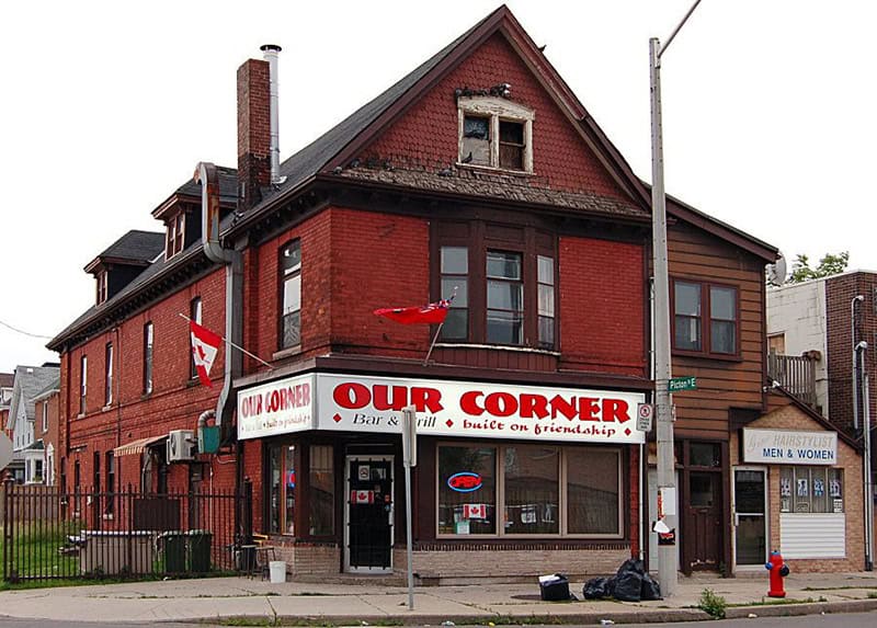 A two-story red brick building houses “OUR CORNER Bar & Grill” on a street corner, with an adjacent restroom and various signs visible. A black trash bag is on the sidewalk, while the welcoming atmosphere inside reflects values inspired by Jesus’ teachings and emphasizes a sense of community rooted in Christian faith.