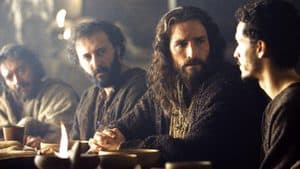 Four men in robes sit at a table with food and drink, engaged in conversation in a dimly lit setting, sharing stories that reflect the bonds of masculinity.