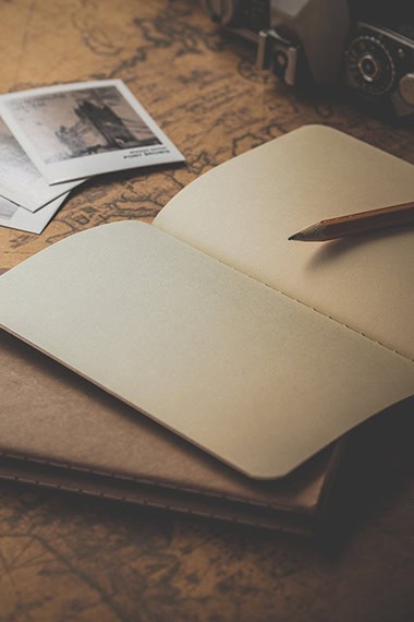 An open blank notebook with a pencil rests on a map, with two old photographs and a vintage camera visible in the background.