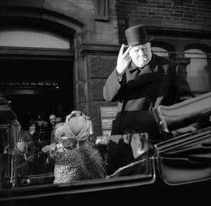 Winston Churchill stands in an open-top car, embodying resilience as he wears a top hat, smokes a cigar, and makes a V-for-victory sign amid hardship.