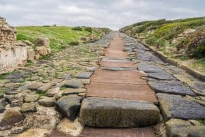 A path of ancient stone and weathered boards cuts through a ruined site, evoking the weight of past suffering across the desolate, grassy landscape.