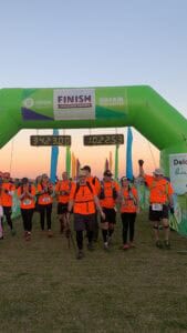 A group of hikers in bright orange shirts cross a green finish line arch for the Oxfam Trailwalker event, showing visible resilience after their long journey.
